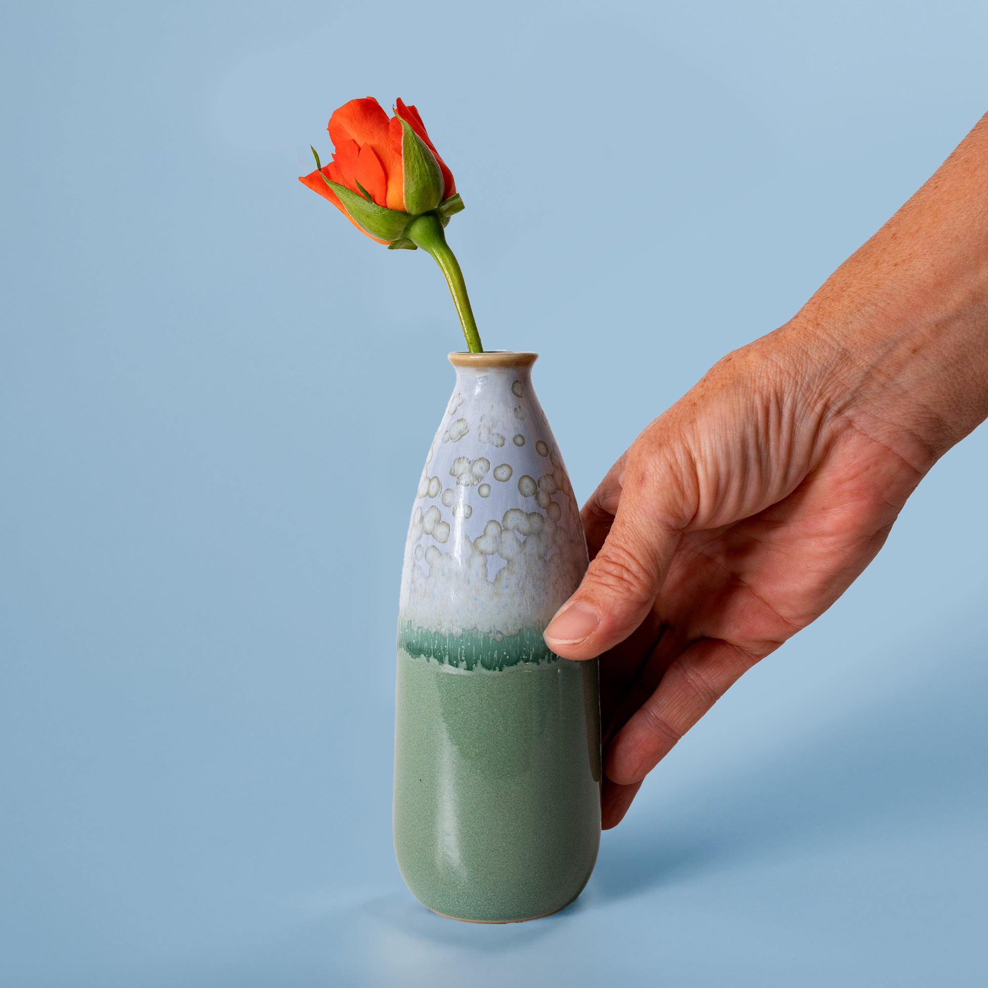Hand holding artisan decorative vase for flowers with green and white glaze and single orange rose