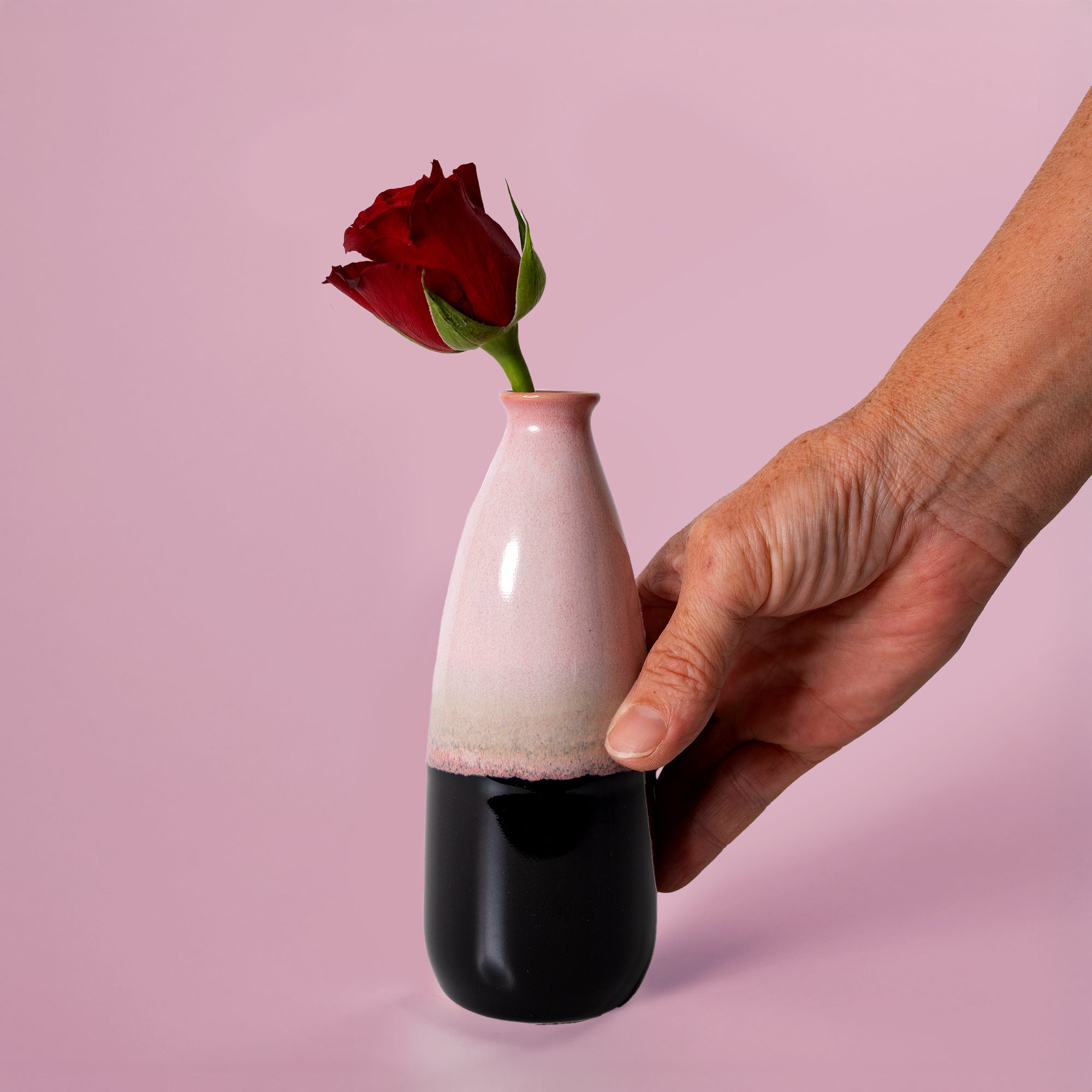 Hand holding an artisan decorative vase for flowers with pink and black glaze and a red rose