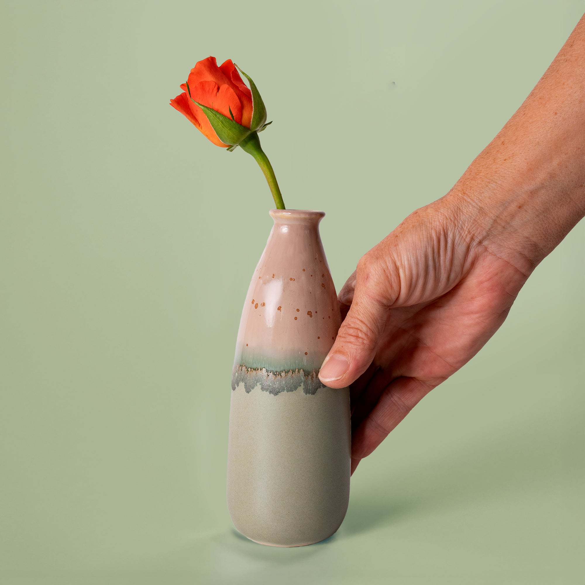 Hand holding artisan decorative vase for flowers with pink and green glaze and single orange rose