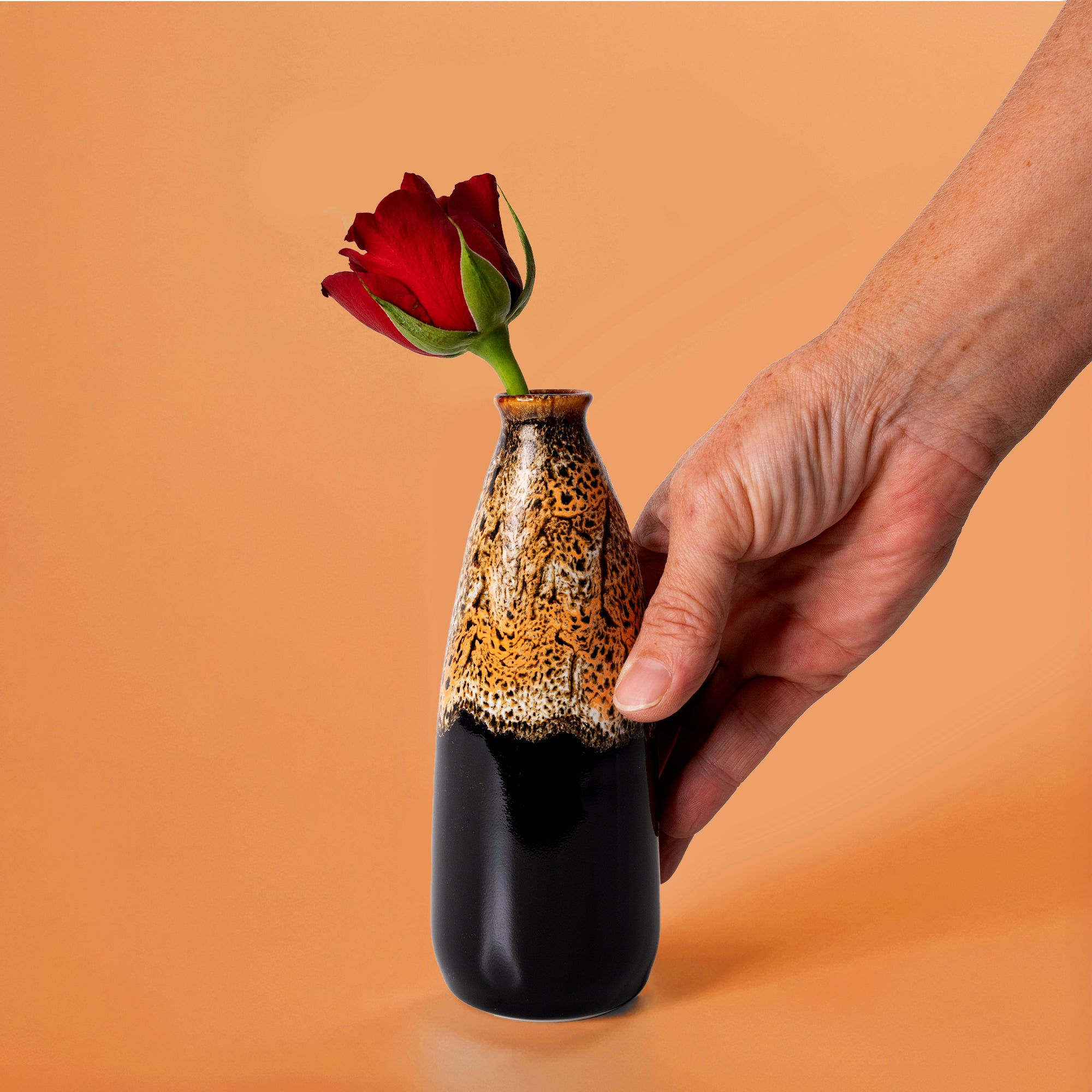 Hand holding artisan decorative vase for flowers with textured brown and black glaze and a red rose