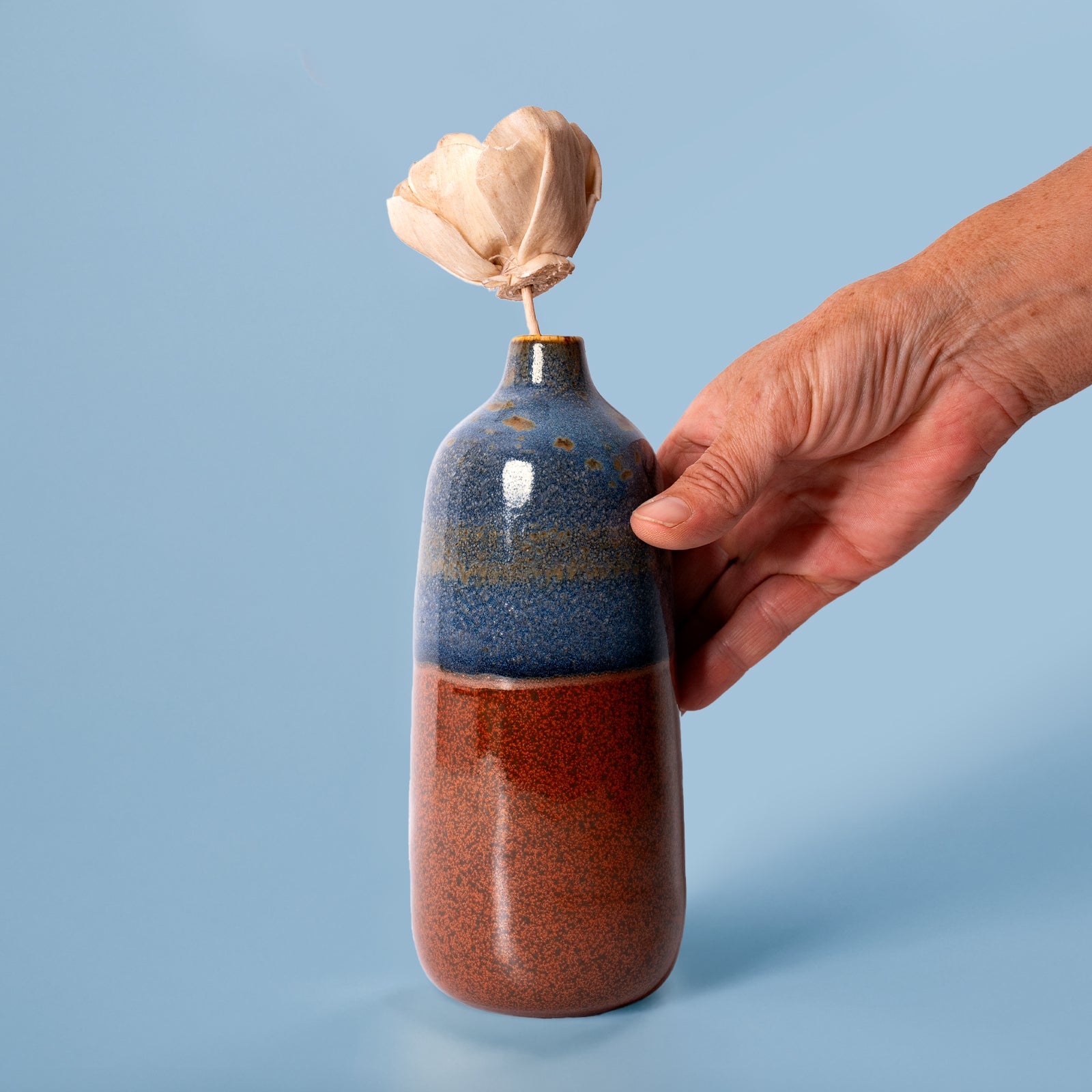 Hand holding Pottery For The Planet eco-friendly home decor vase with blue and brown glaze and dried flower
