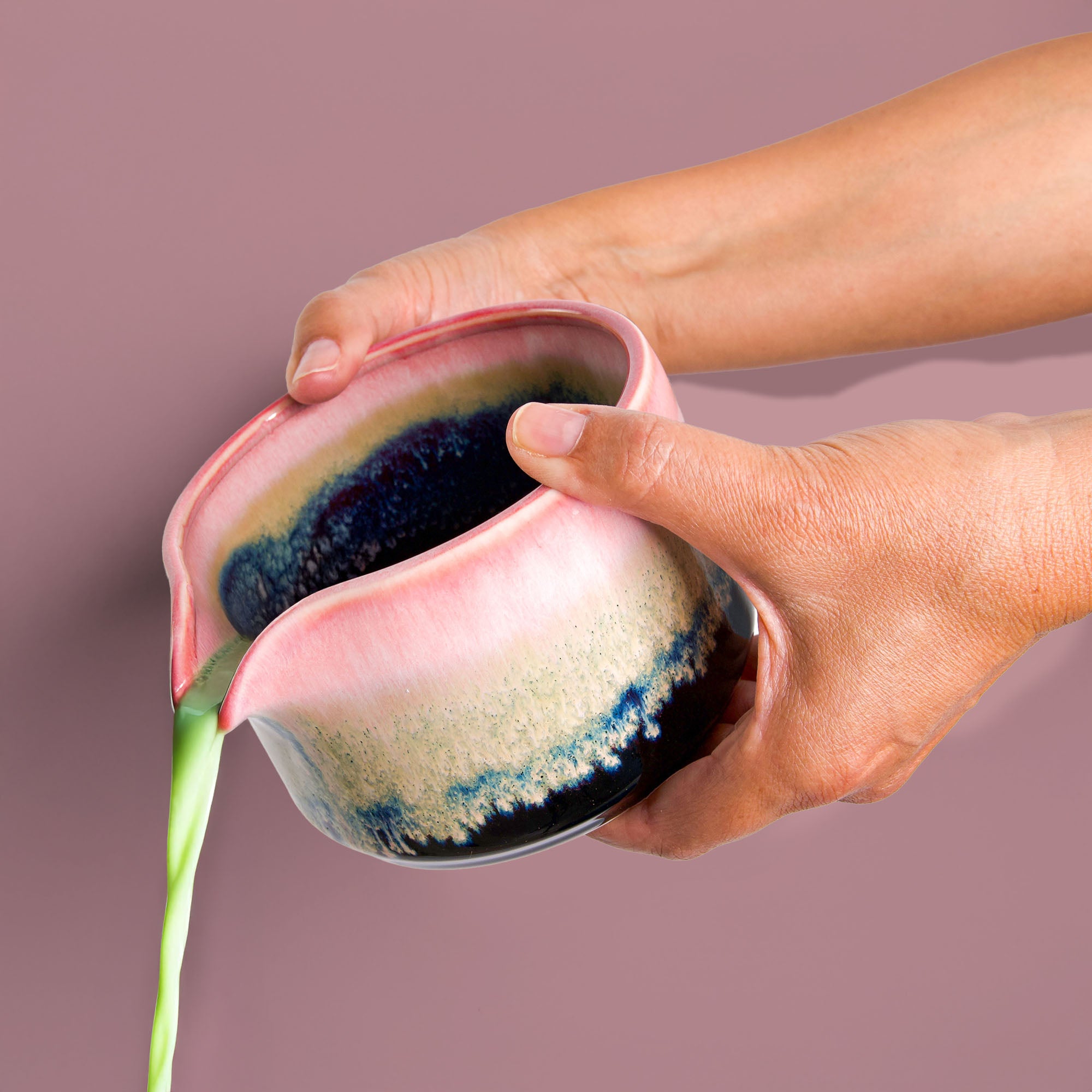 Hand pouring green tea from a handcrafted matcha bowl with pink and blue glaze by Pottery For The Planet