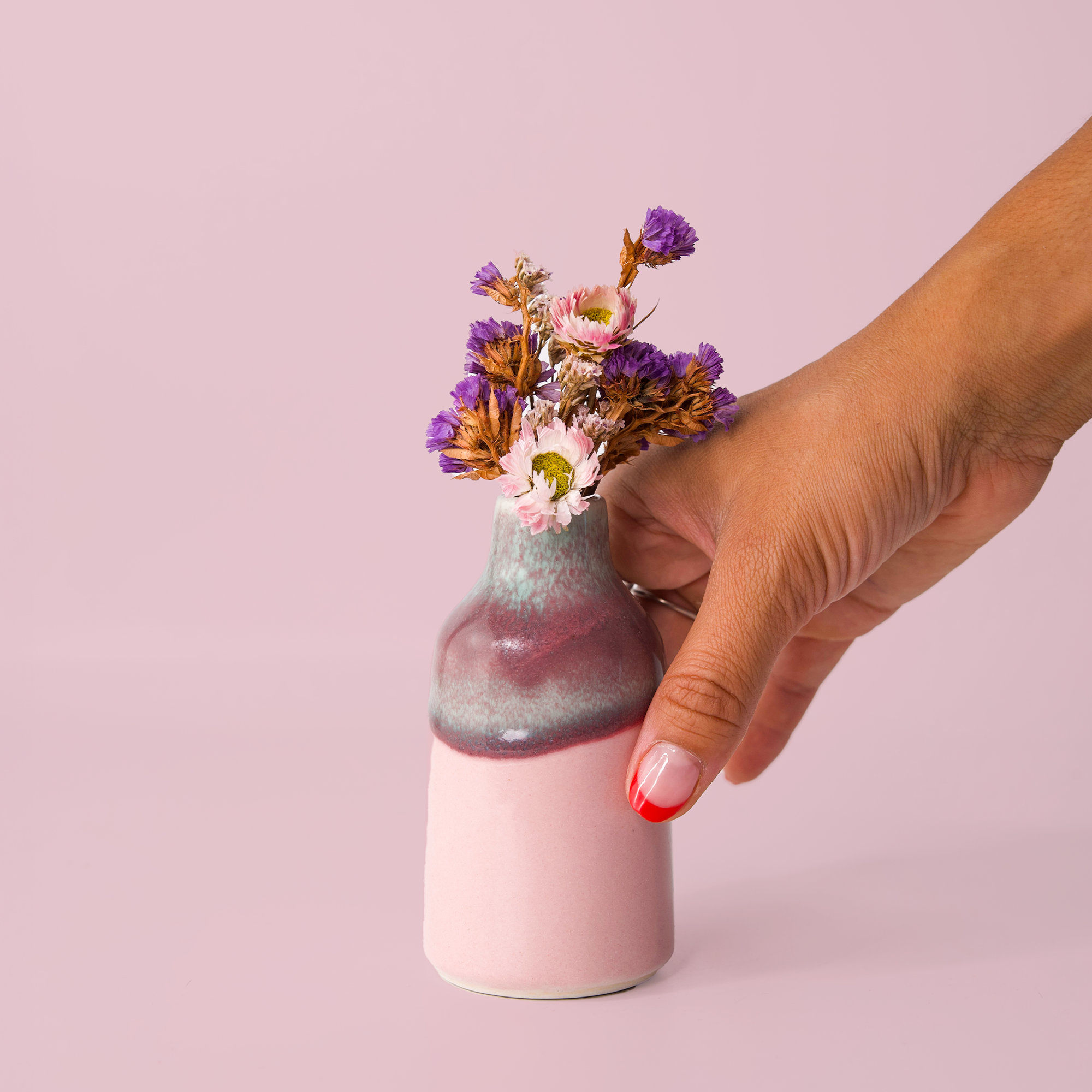 Hand holding modern decorative flower vase with purple and pink dried flowers on pink background