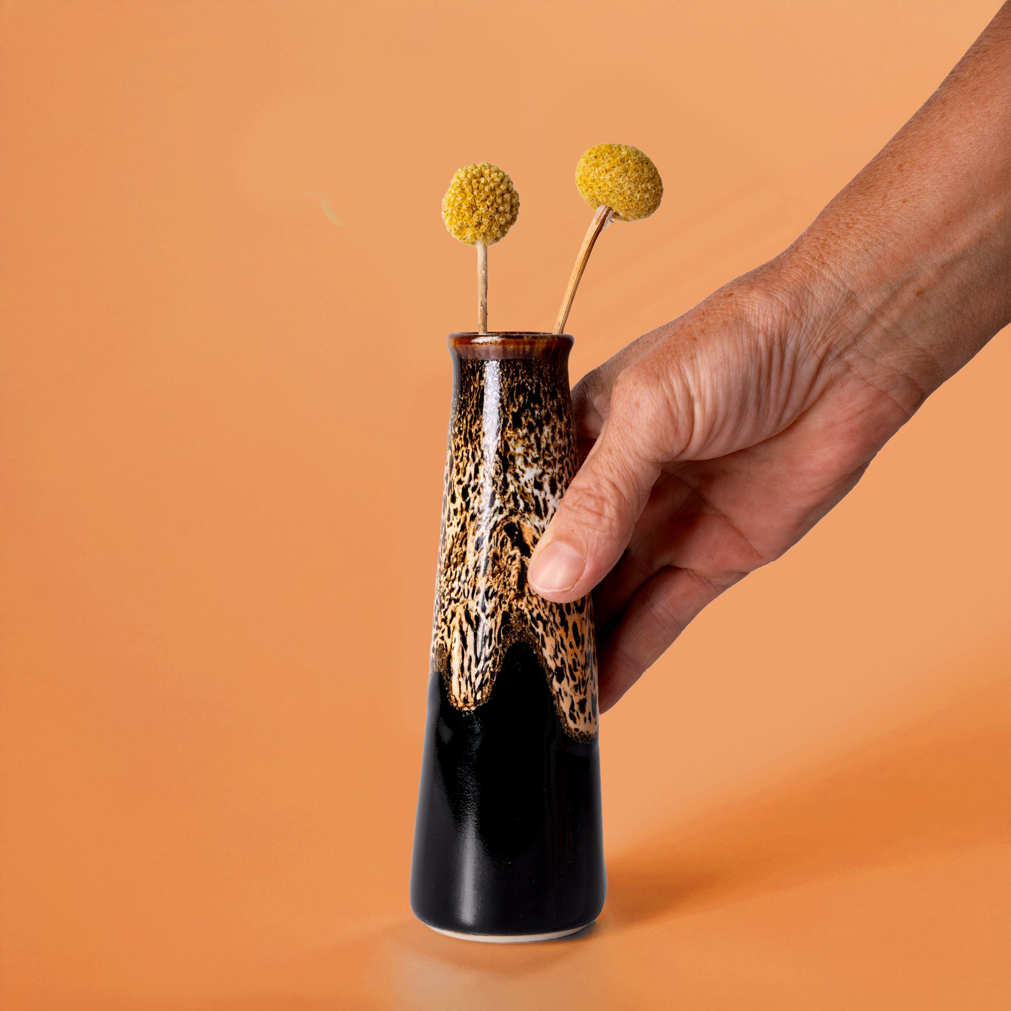 Hand holding Pottery For The Planet sustainable home decor vase with brown and black glaze and yellow billy ball flowers