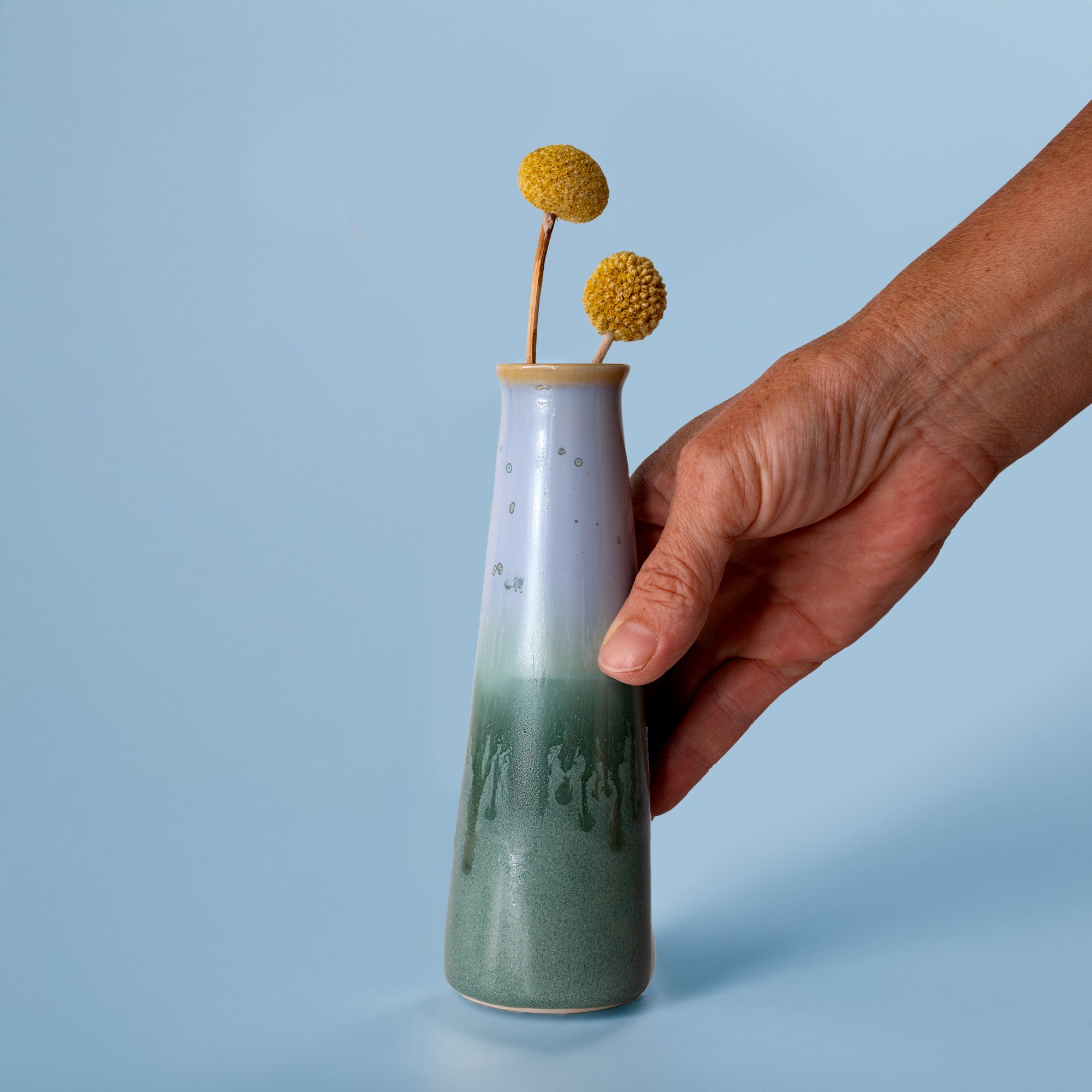 Pottery For The Planet sustainable home decor vase with green and white glaze held by hand with dried yellow flowers