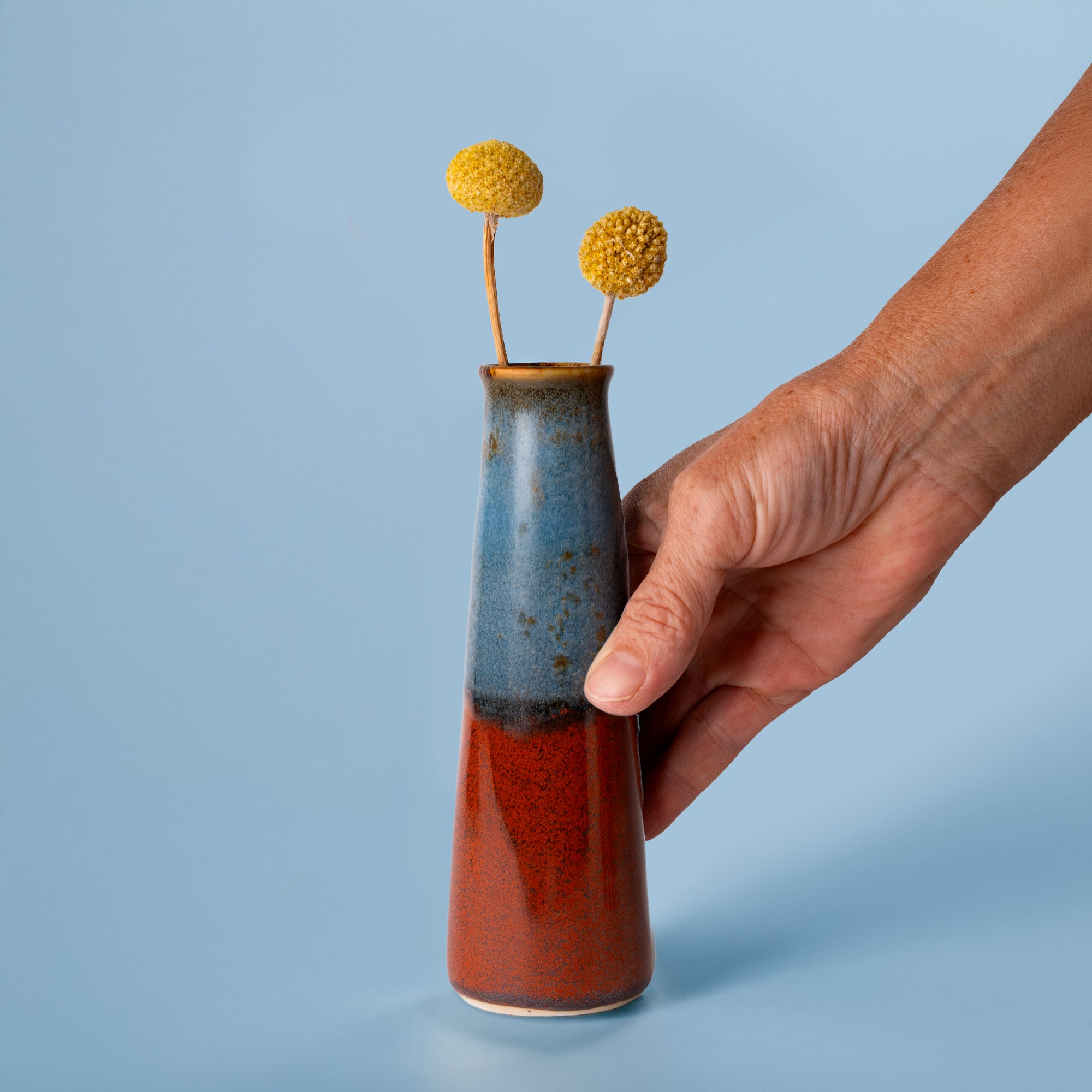 Hand holding a red and blue sustainable home decor vase with two yellow dried flowers by Pottery For The Planet