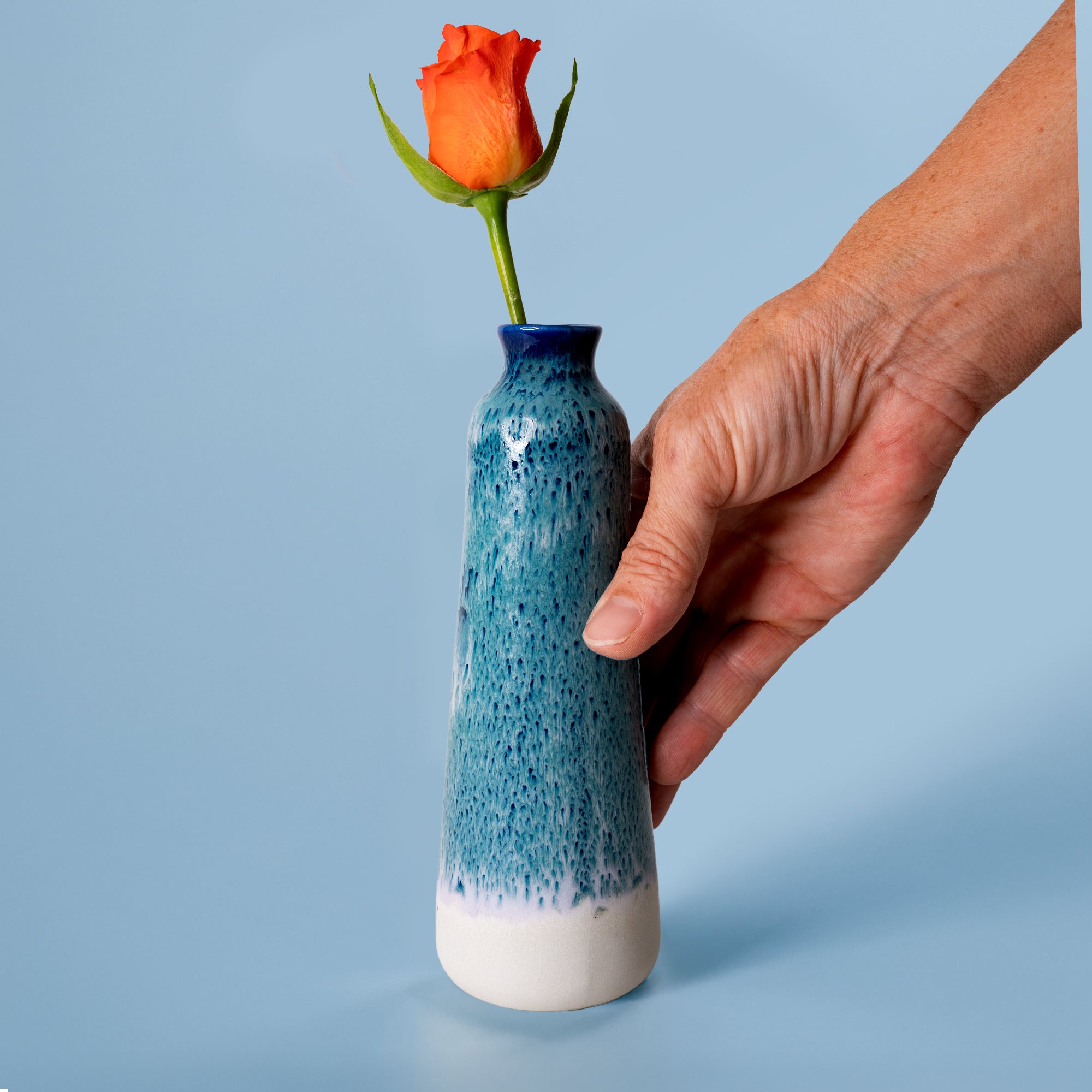 Hand holding unique handmade ceramic vase with blue speckled glaze and orange rose by Pottery For The Planet