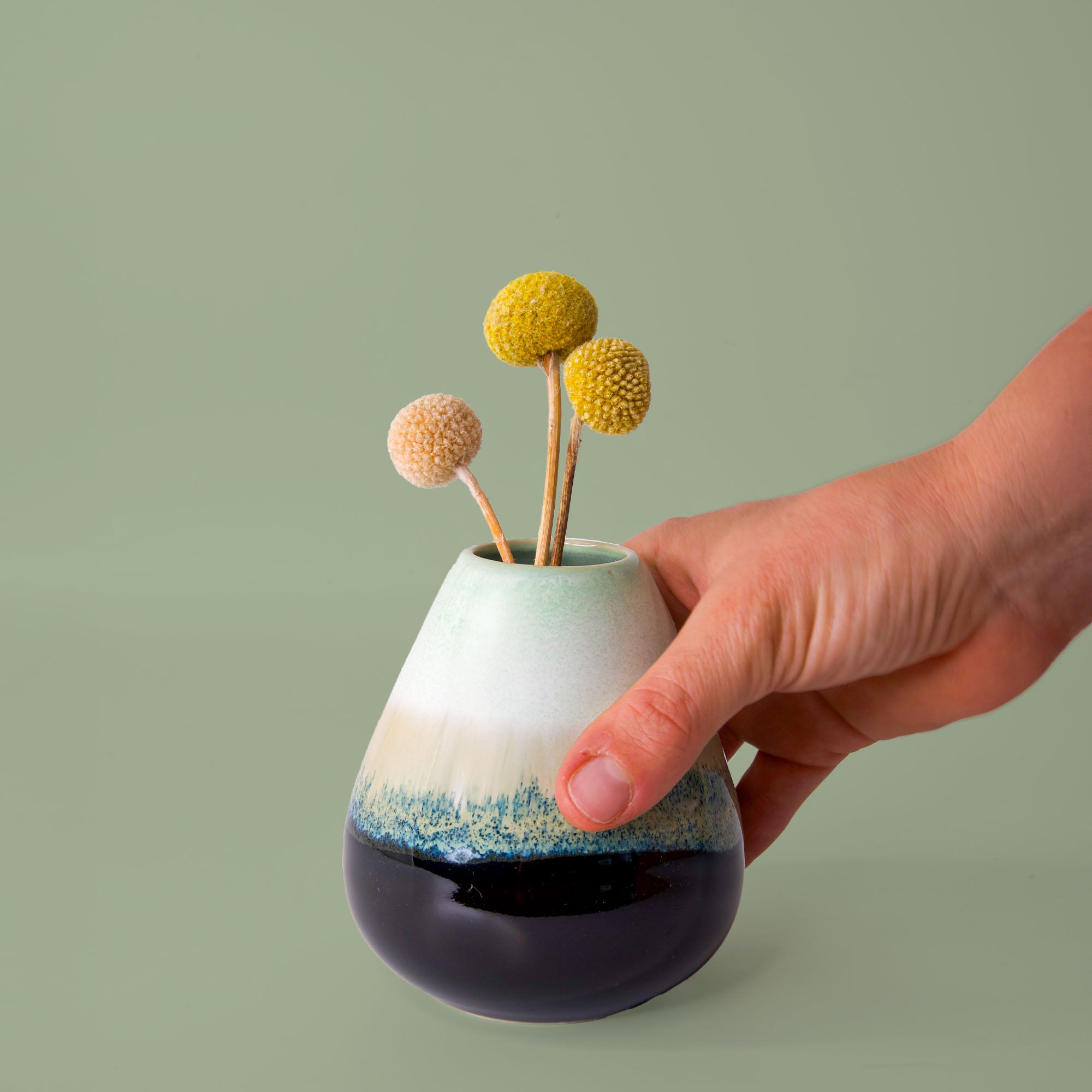 Hand holding unique pottery flower vase with blue, white, and green glaze with dried yellow and peach flowers