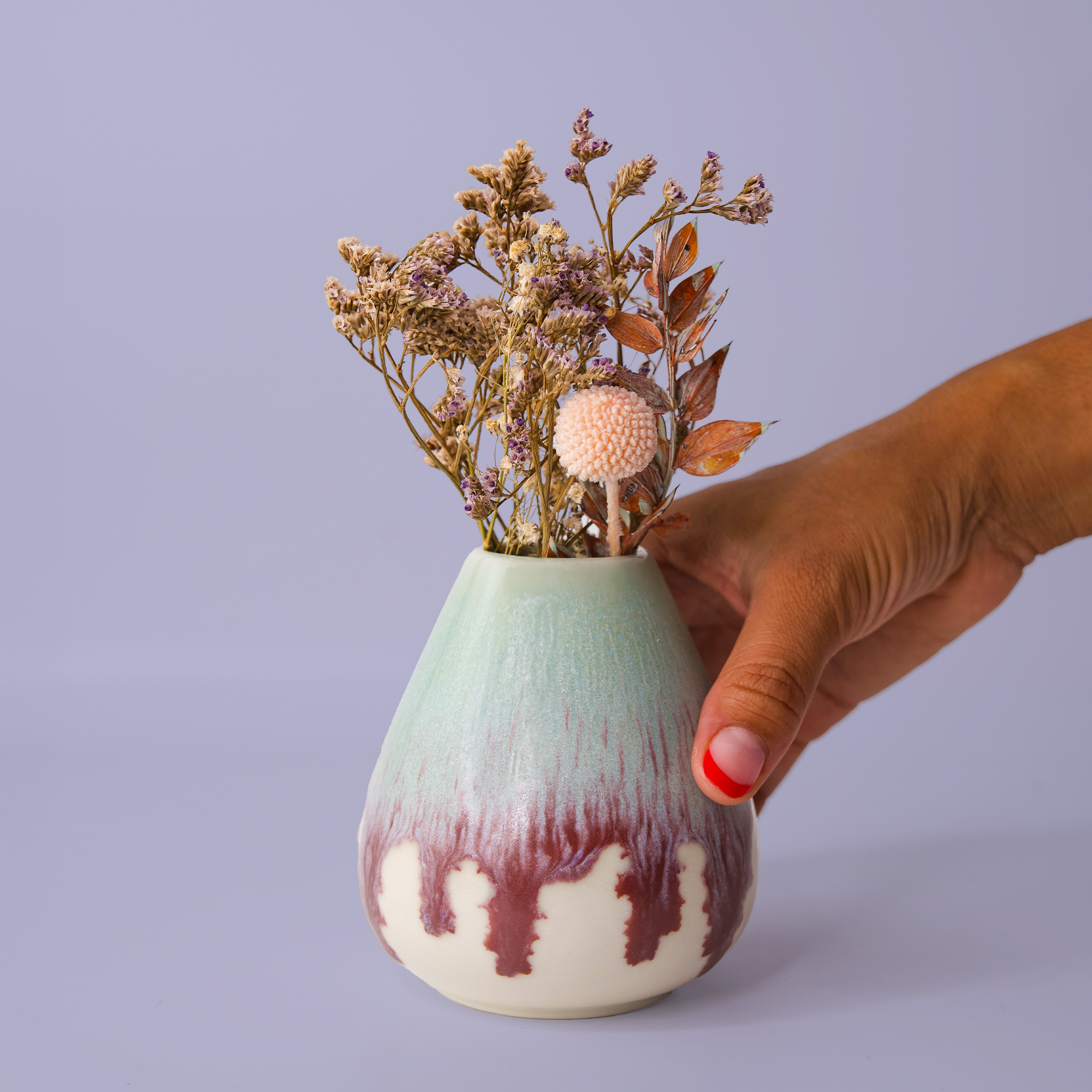 Hand holding a unique pottery flower vase with dried flowers in pastel green and maroon glaze