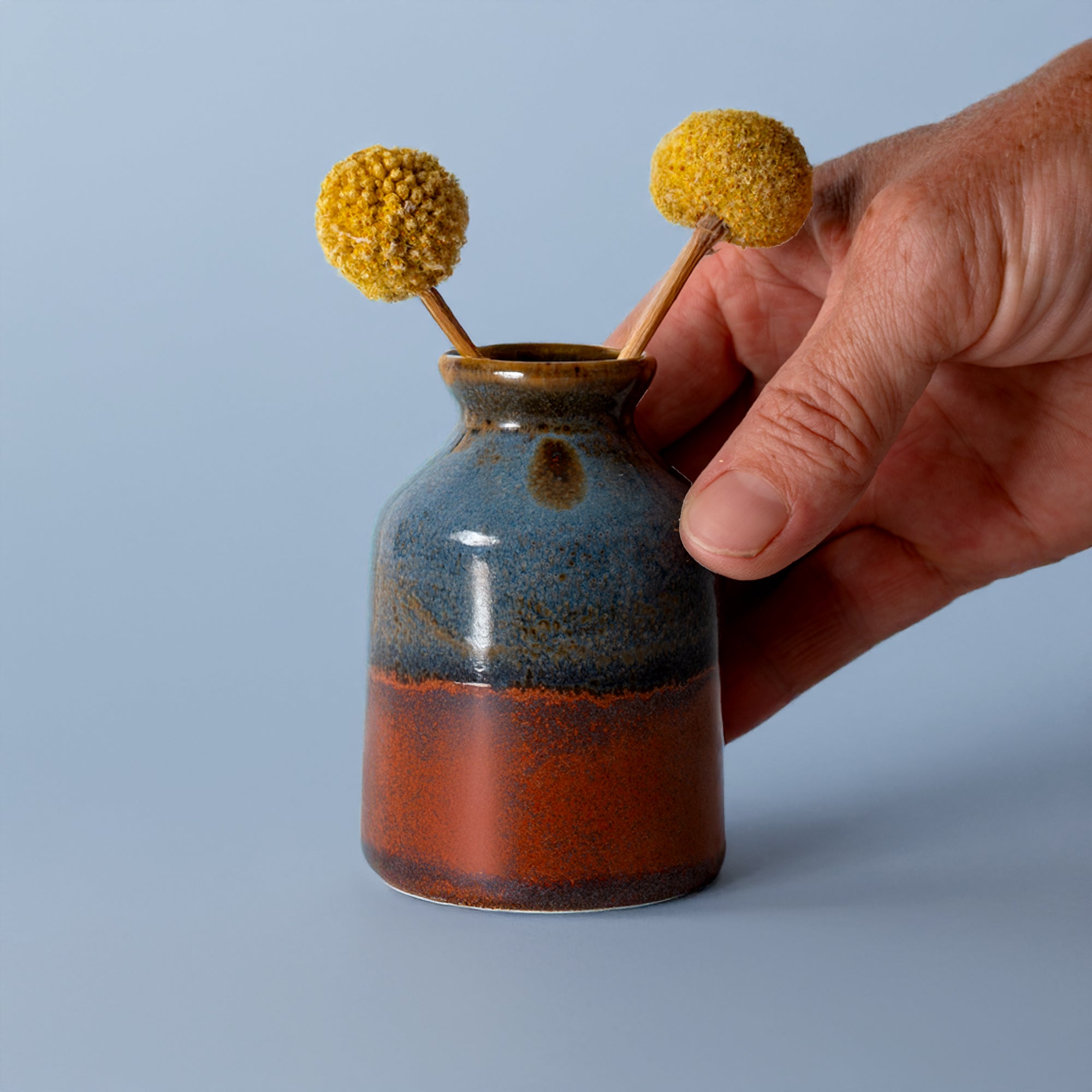 Hand holding a miniature decorative vase for shelf with blue and brown glaze containing yellow billy ball flowers