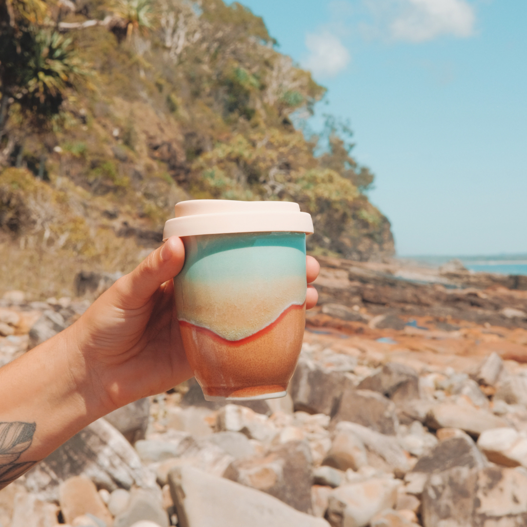 Hand holding a colorful handmade reusable cup by Pottery For The Planet outdoors near rocky shore
