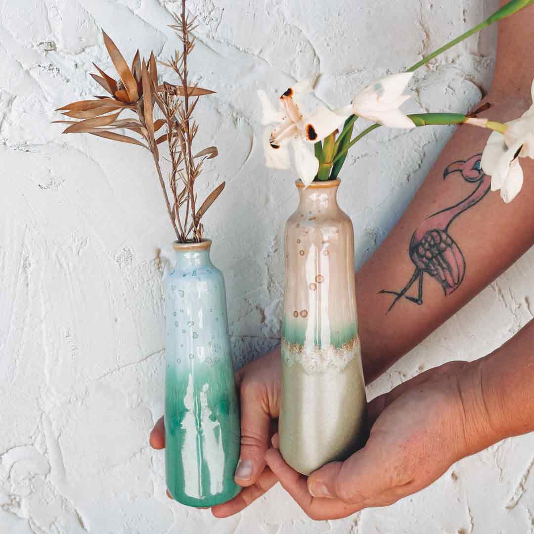 Two unique handmade ceramic vase pieces with gradient glaze held by hands against textured wall background