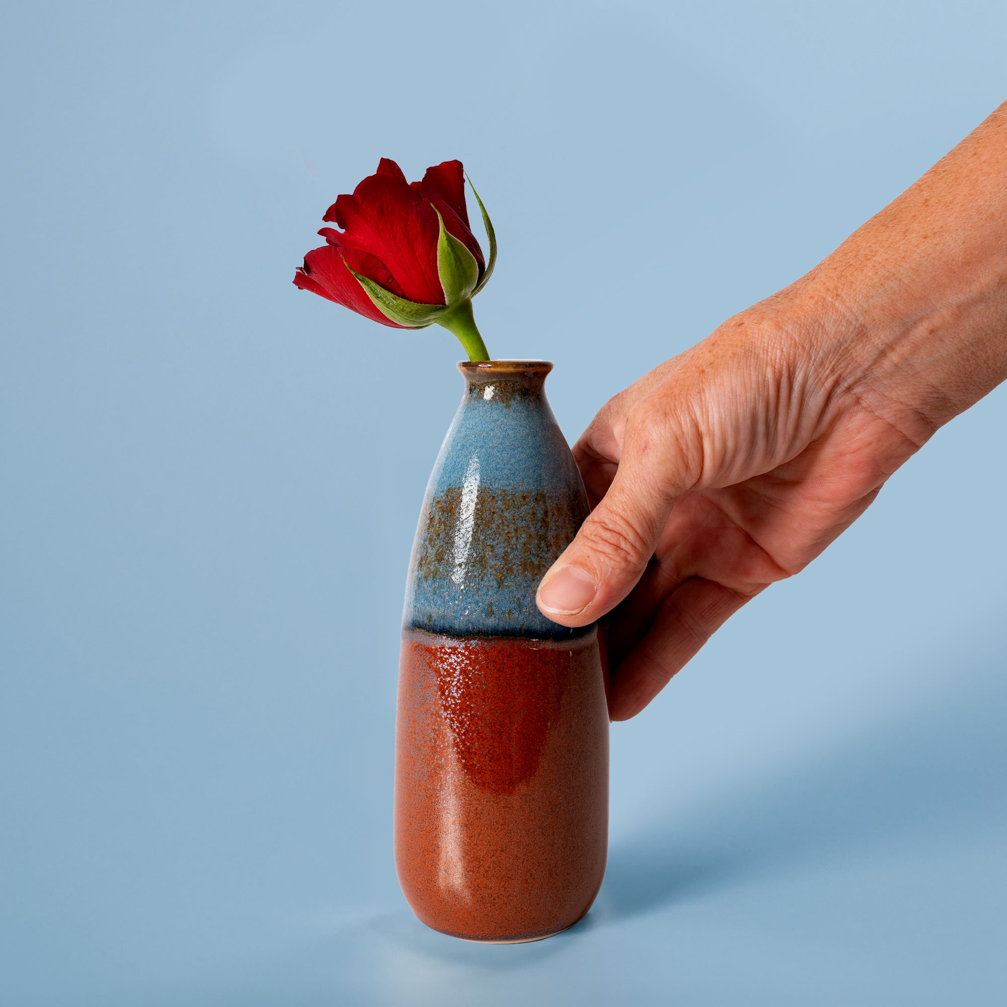 Hand holding artisan decorative vase for flowers with blue and brown glaze and a red rose