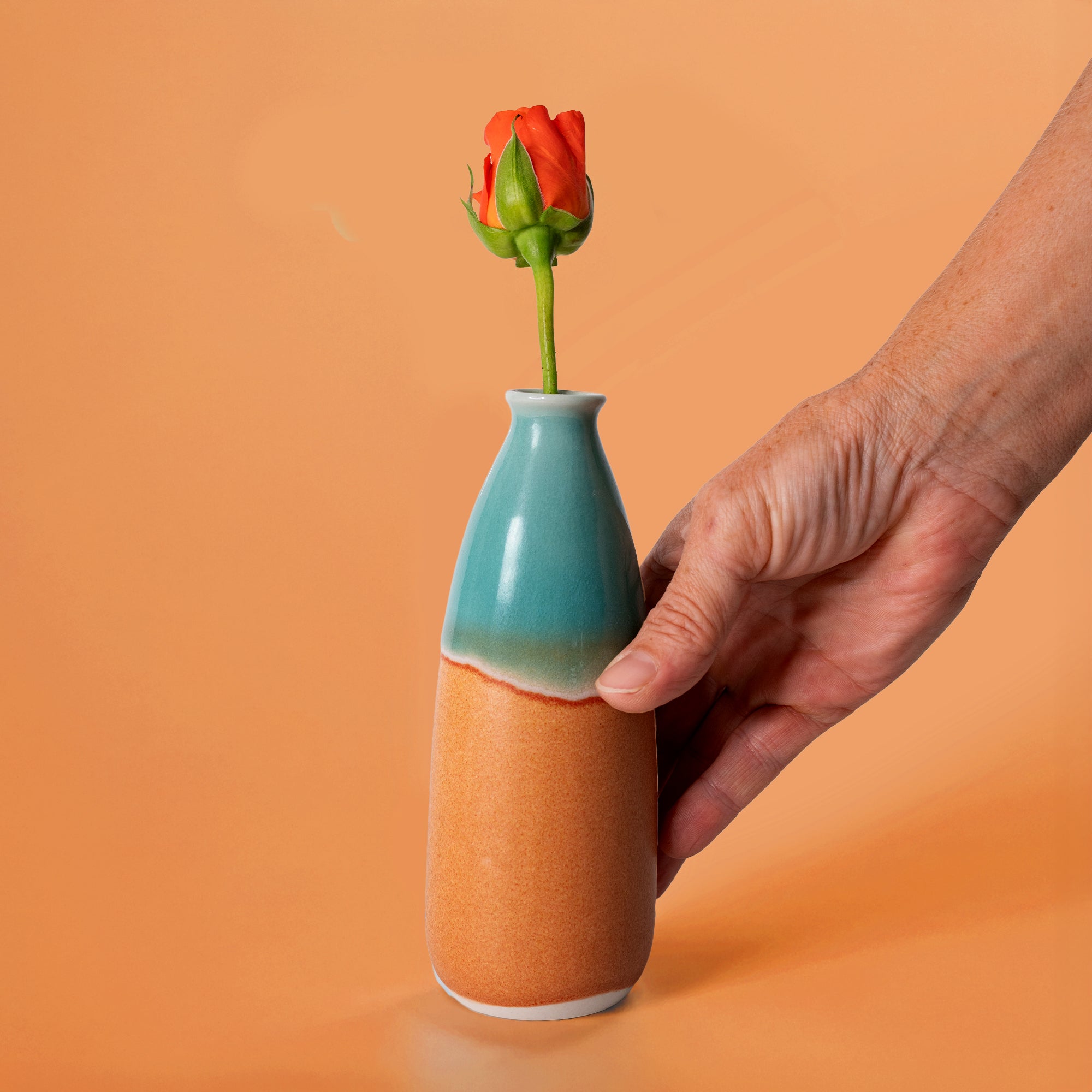 Hand holding a two-tone artisan decorative vase for flowers with a single orange rose bud