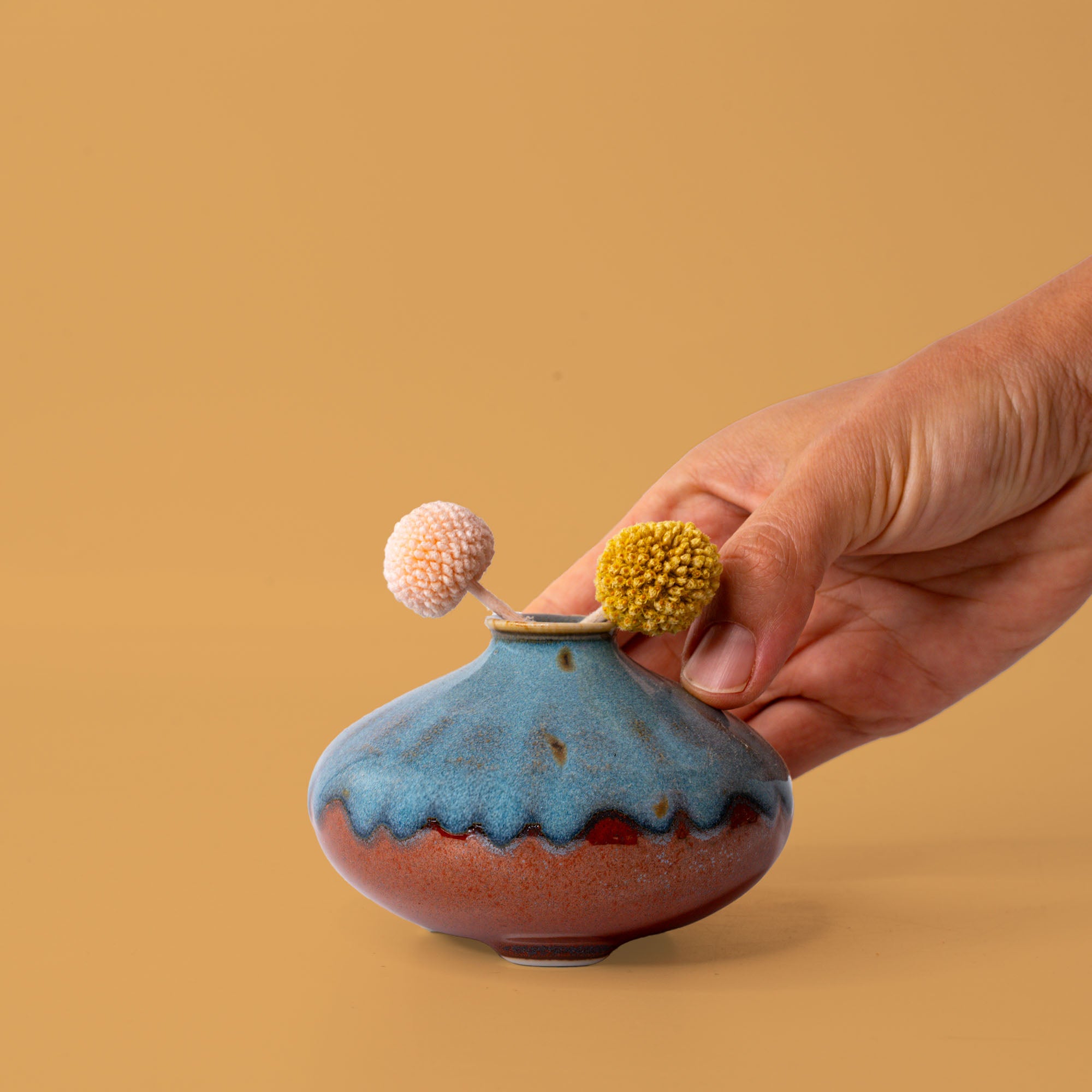 Hand holding a blue and brown artisan decorative vase with two small textured flowers inside