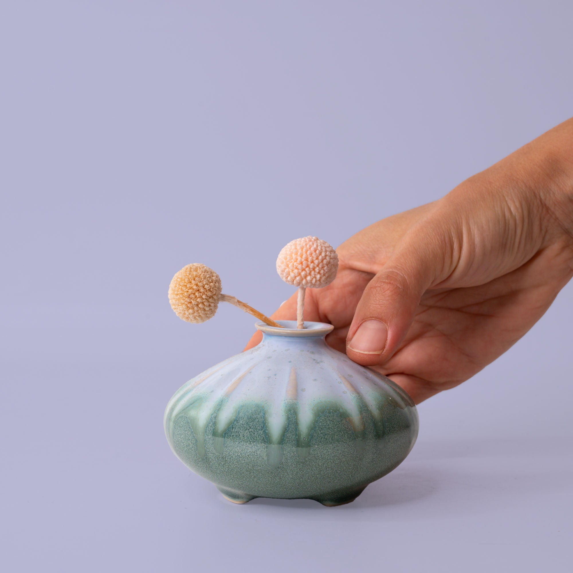 Hand holding a green and white ceramic artisan decorative vase with small dried flowers