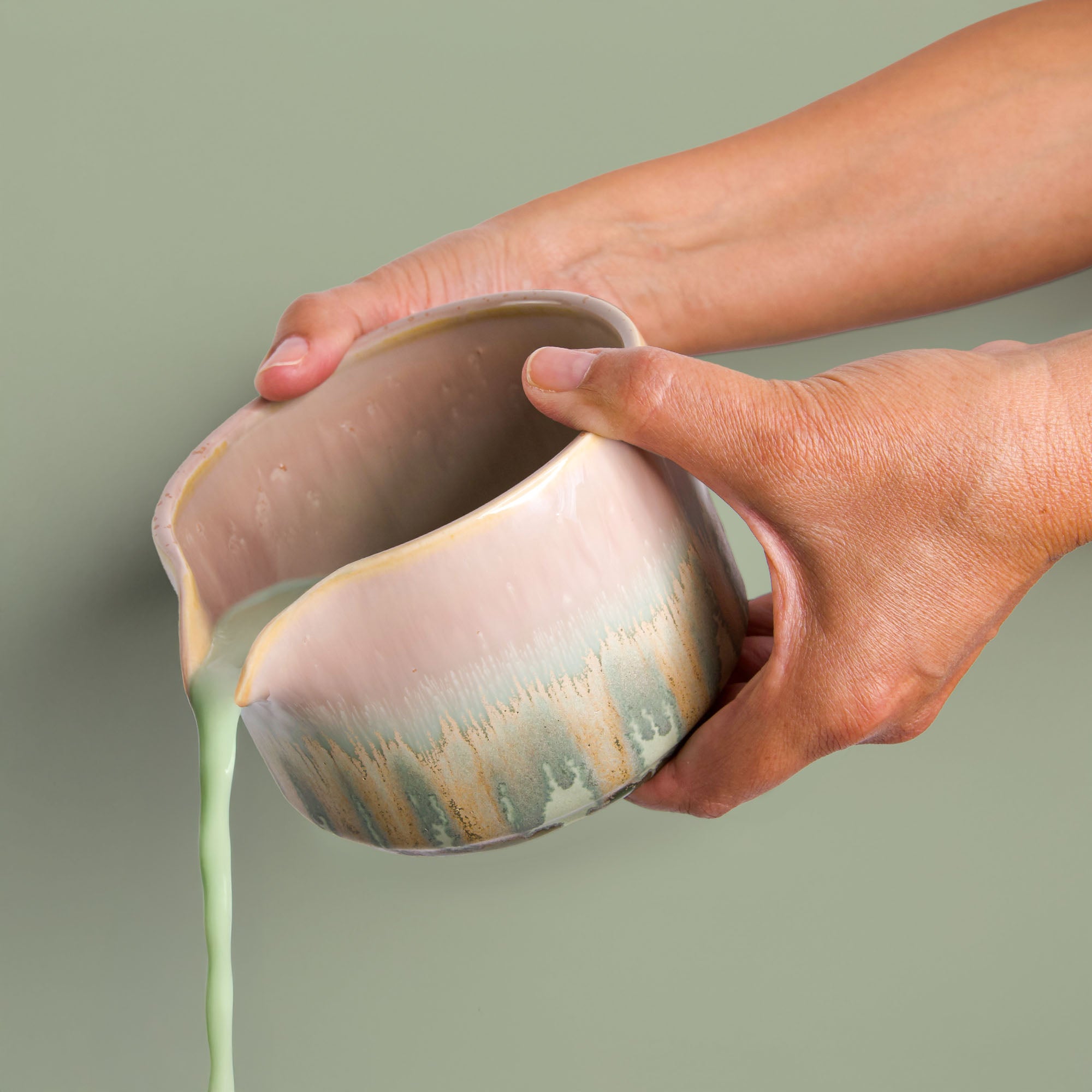 Hand holding a handcrafted matcha bowl pouring green matcha tea against a neutral background