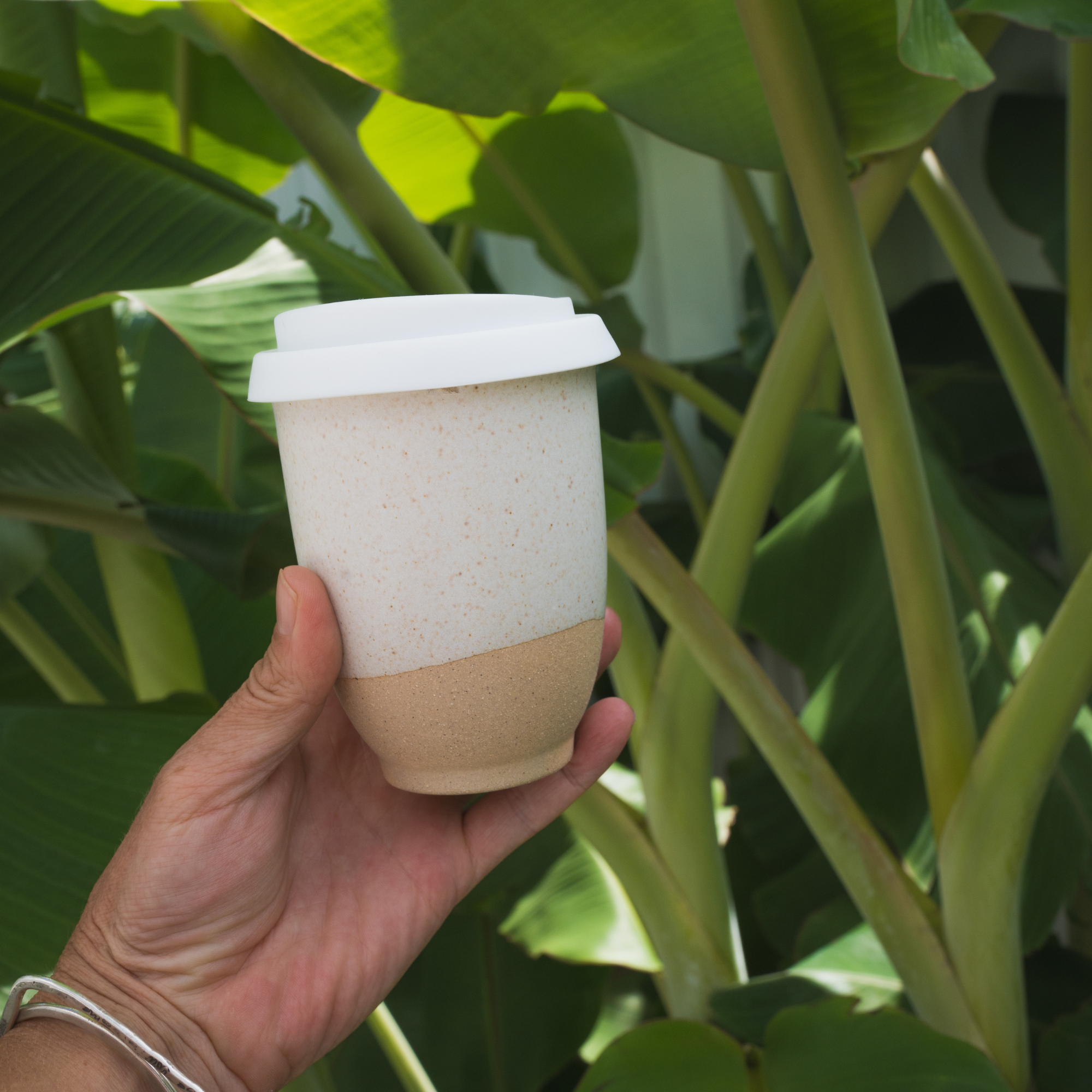 Hand holding a handmade reusable coffee cup with a white lid against green plant background