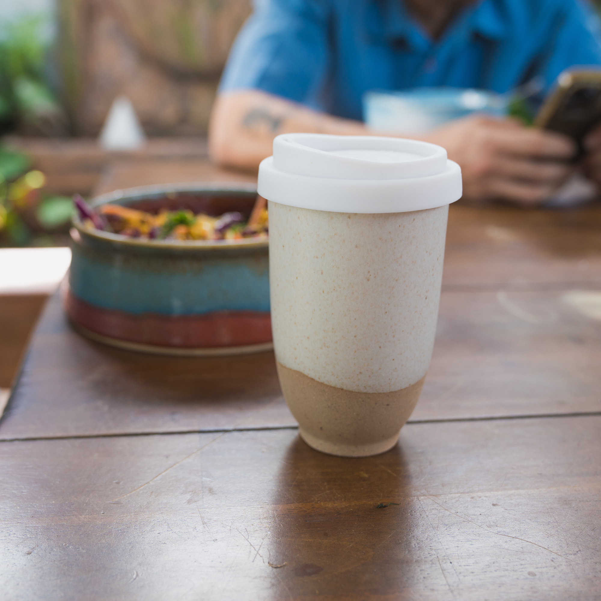 Handmade reusable coffee cup by Pottery For The Planet with white lid on wooden table