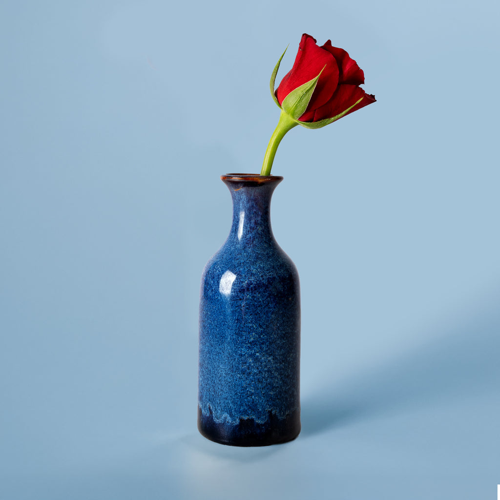 Small handmade flower vase in deep blue ceramic with a single red rose on a light blue background