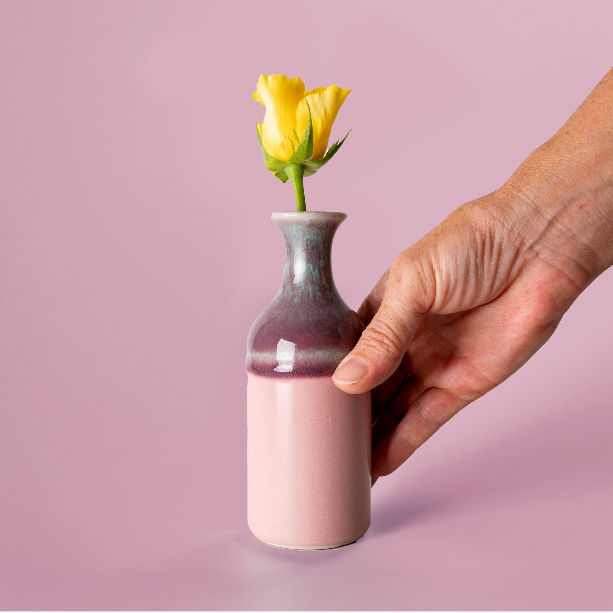 Small handmade flower vase by Pottery For The Planet holding a single yellow rose with hand on pink background
