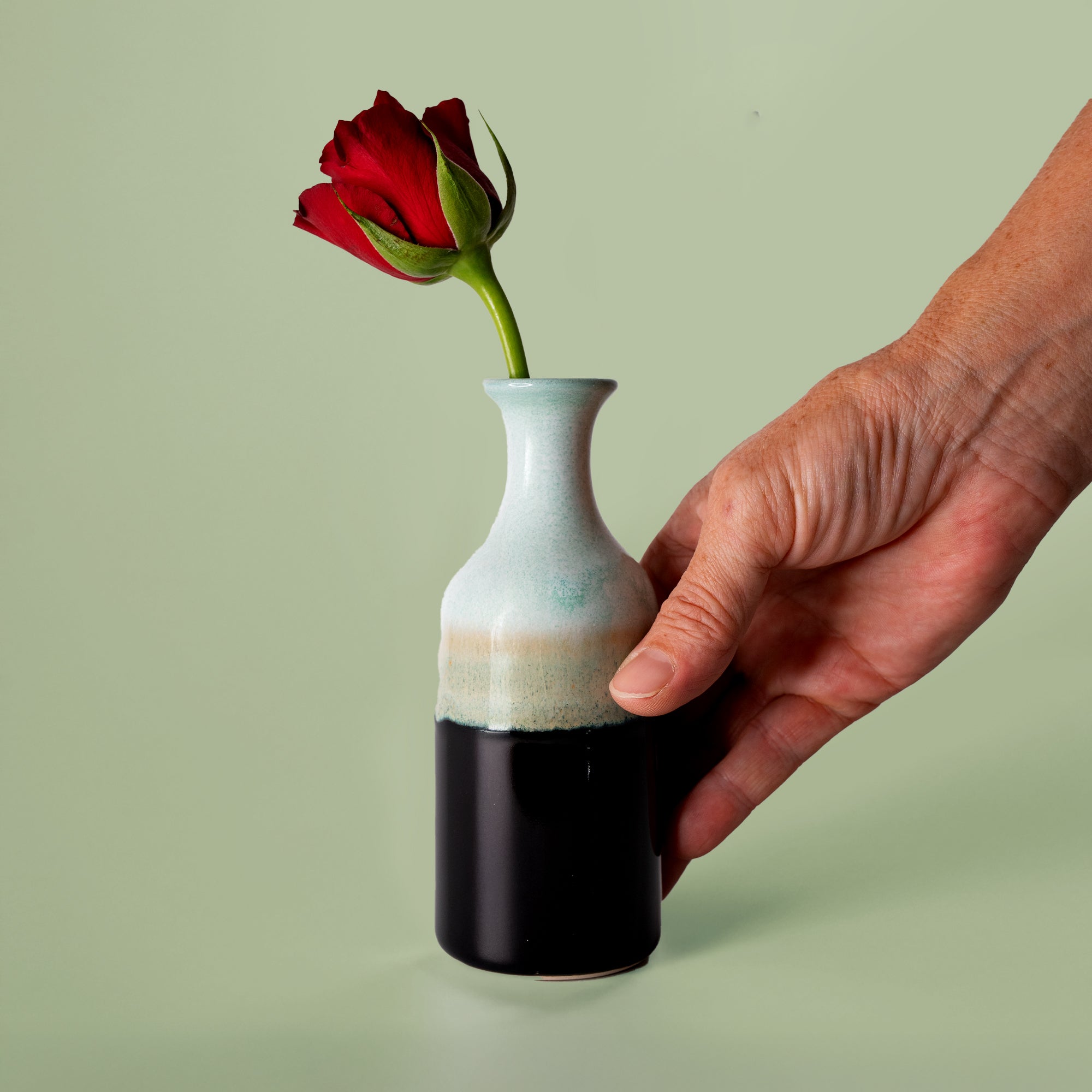 Small handmade flower vase by Pottery For The Planet held with a red rose on green background