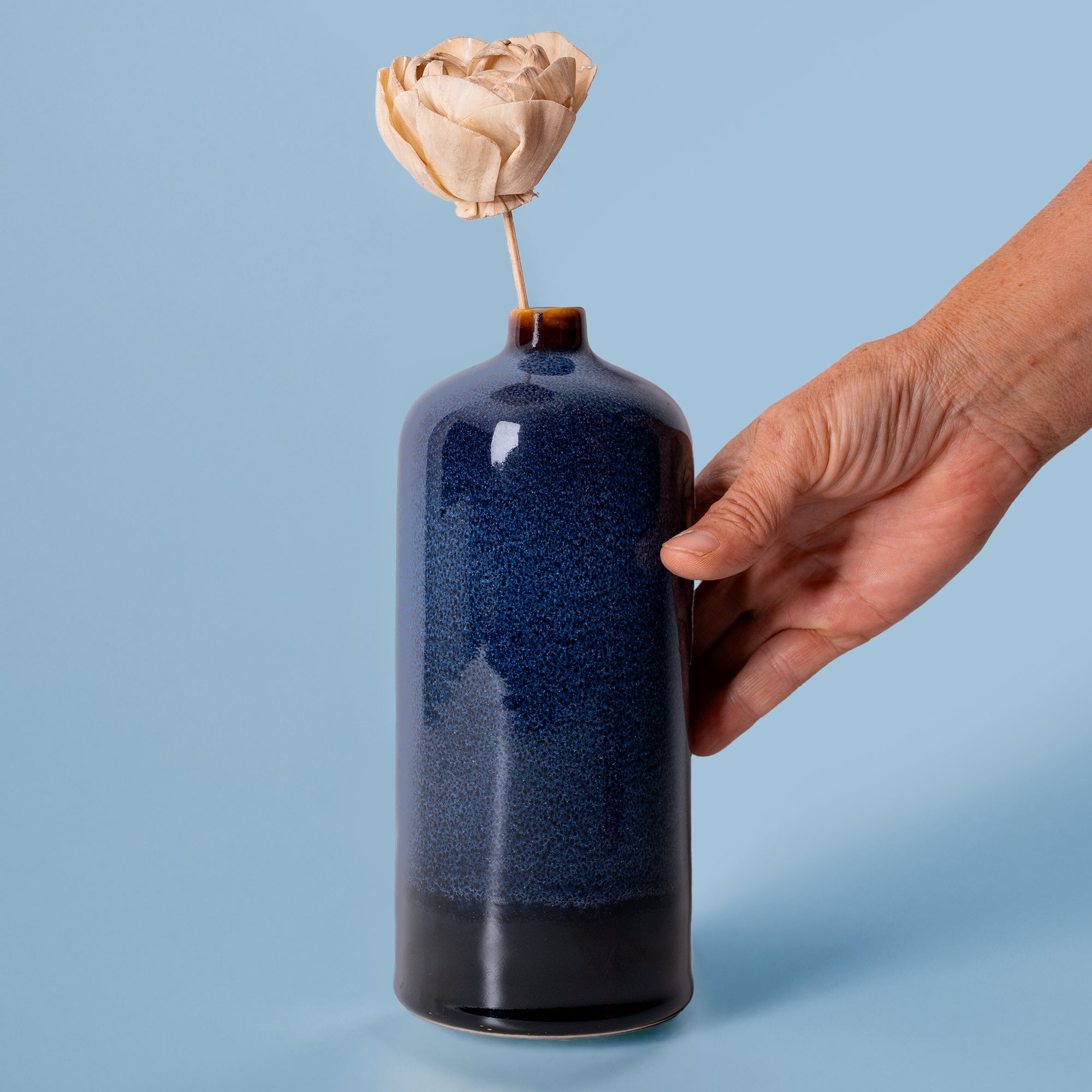 Hand holding a blue ceramic medium decorative bud vase with a single dried flower from Pottery For The Planet