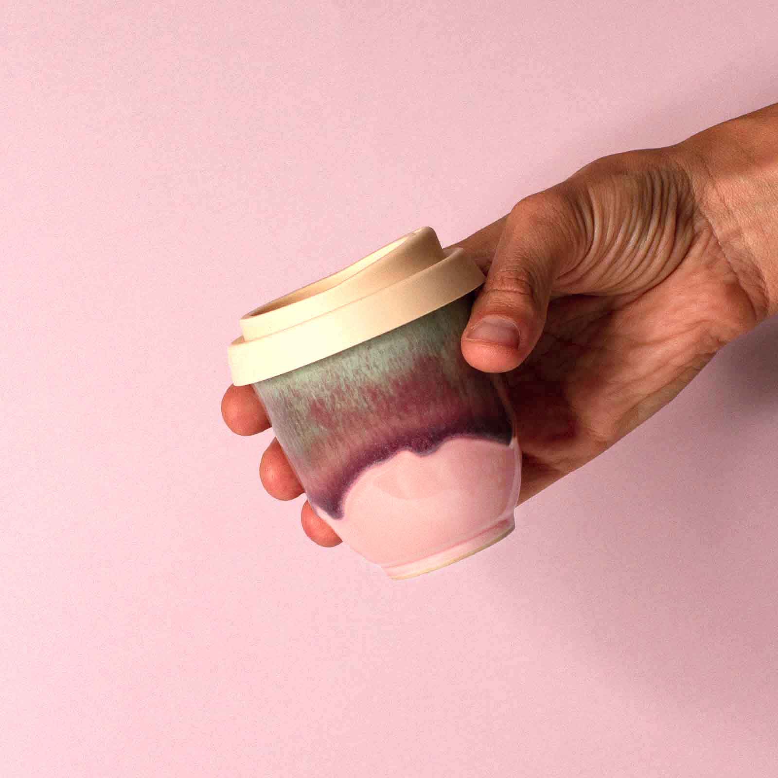 Hand holding pink ceramic travel cup with white lid by Pottery For The Planet on pink background