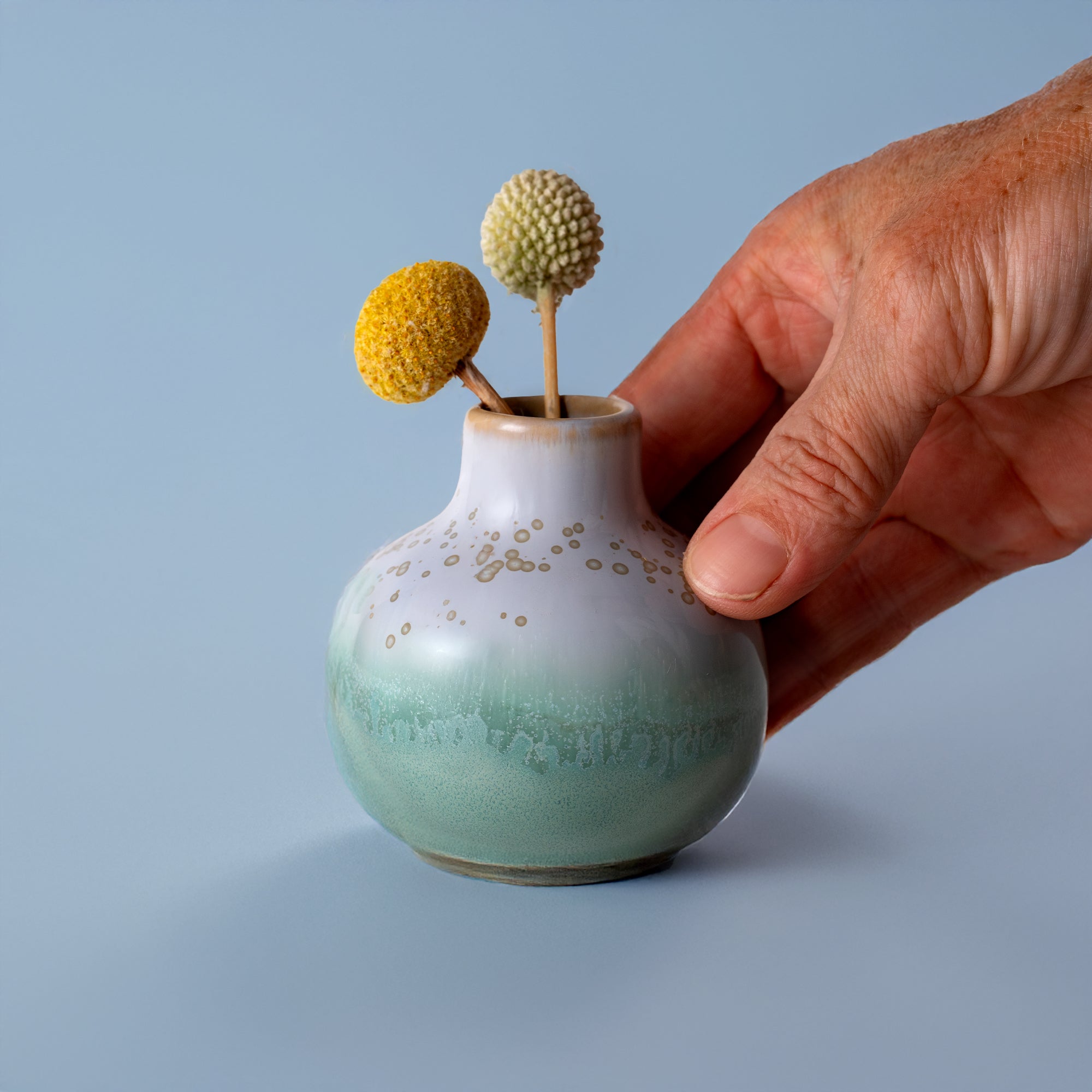 Small handmade home decor vase in green and white glaze held by hand with dried yellow and white flowers