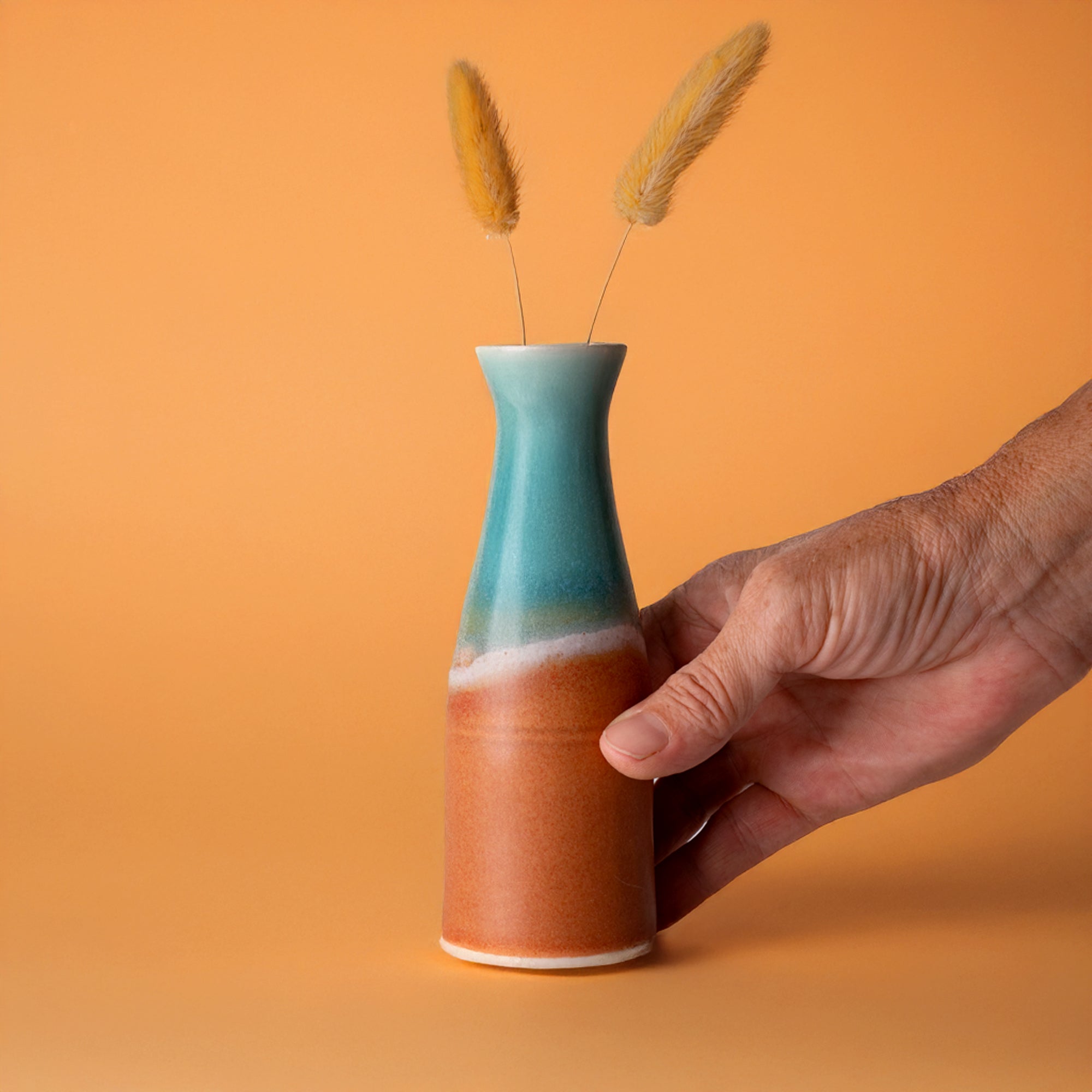 Hand holding a unique ceramic flower vase with blue and brown gradient glaze and dried grass stems