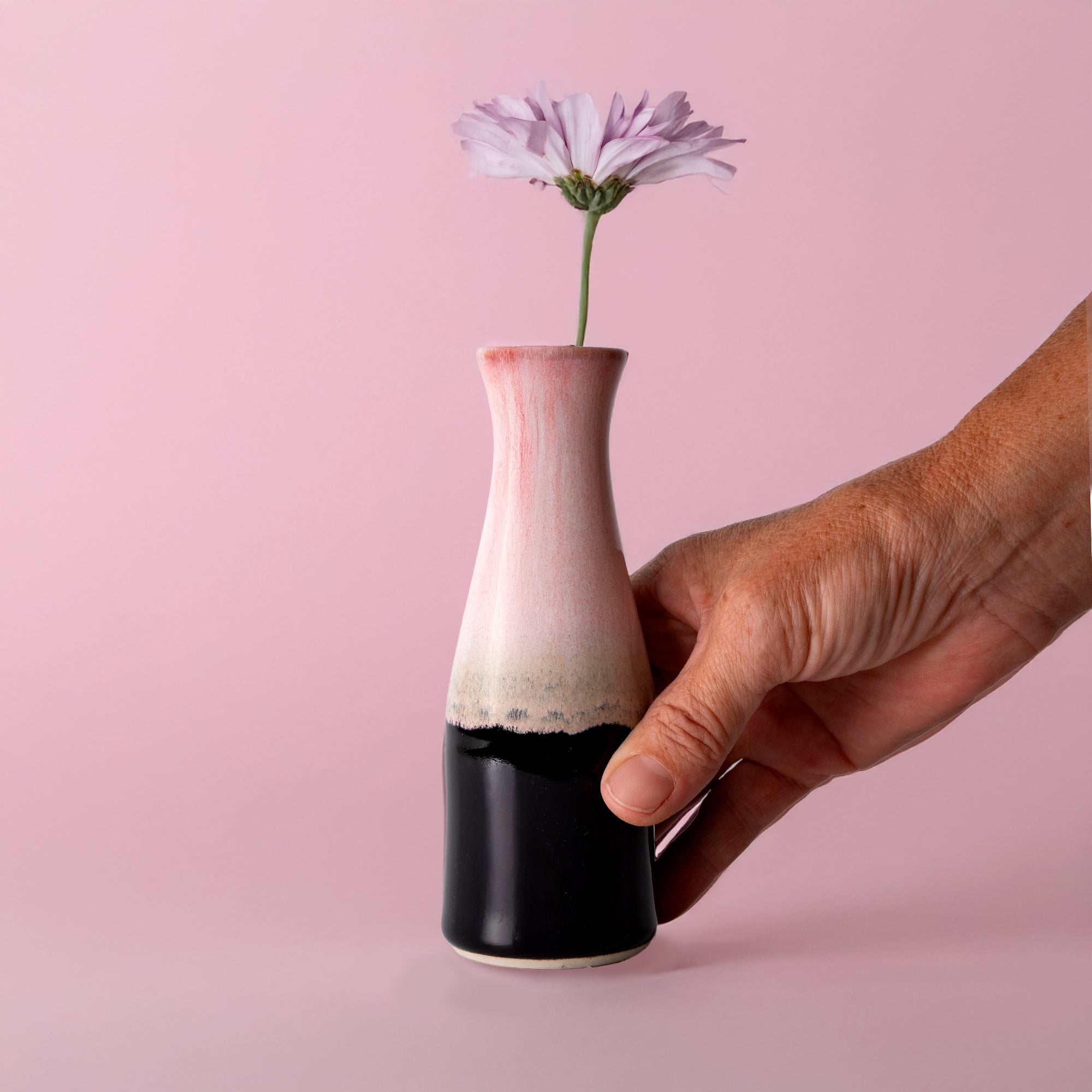 Hand holding a unique ceramic flower vase in pink and black with a single purple flower by Pottery For The Planet