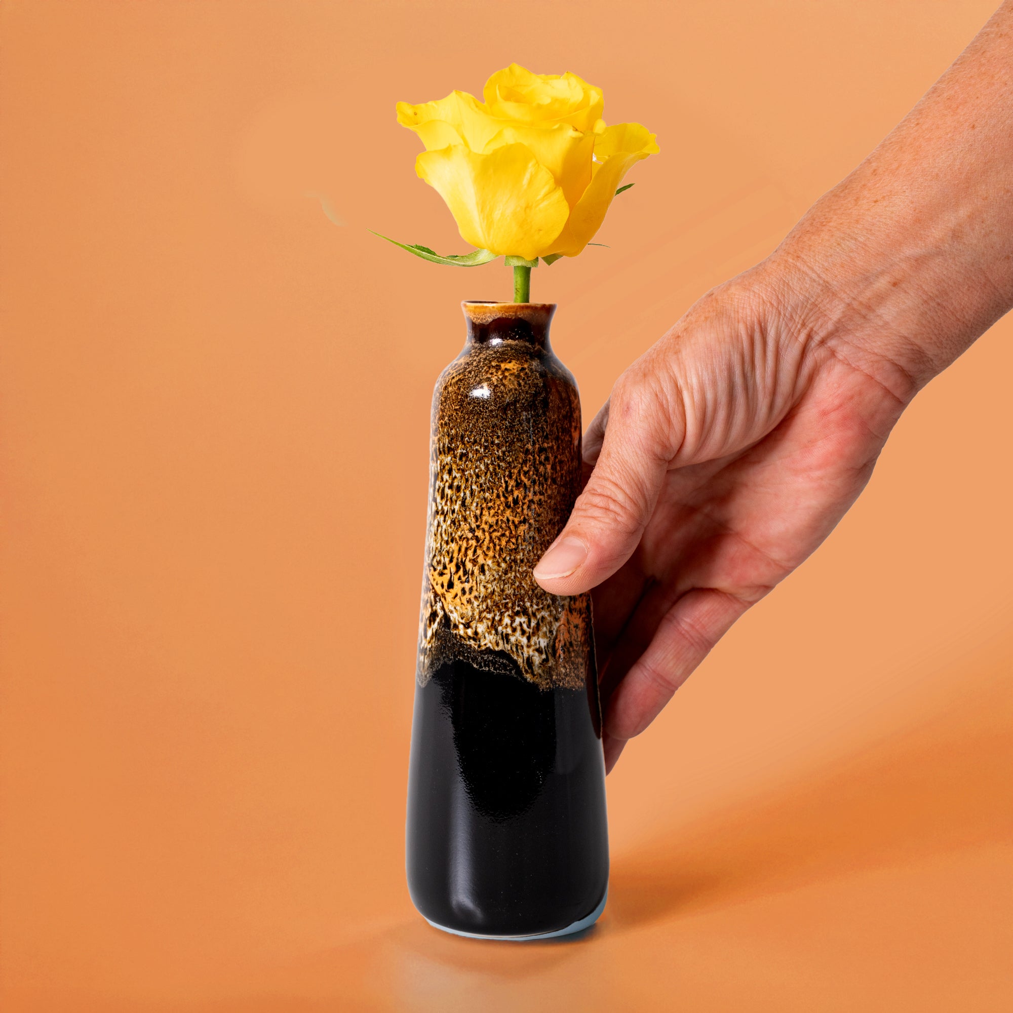 Hand holding a unique handmade ceramic vase with brown and black glaze containing a yellow rose
