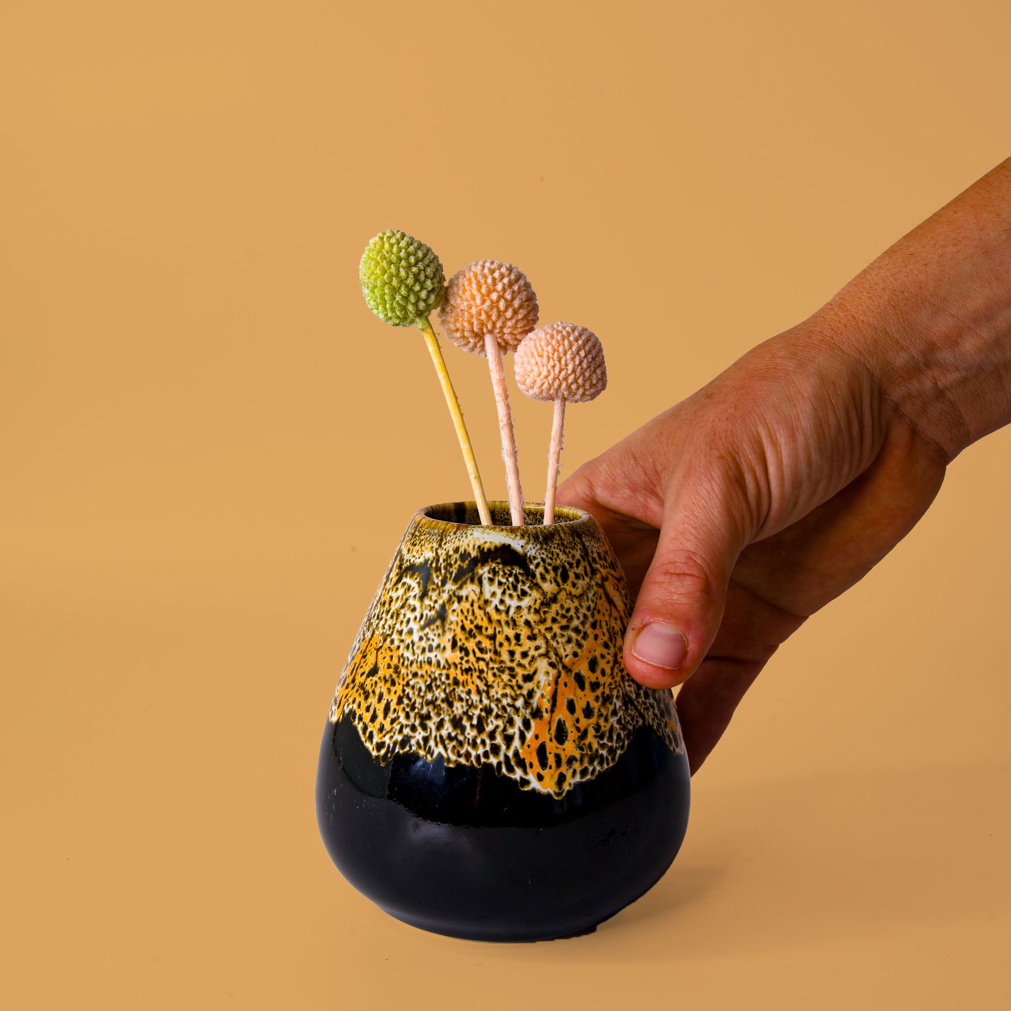 Hand holding unique pottery flower vase with black and yellow speckled design and decorative dried flowers