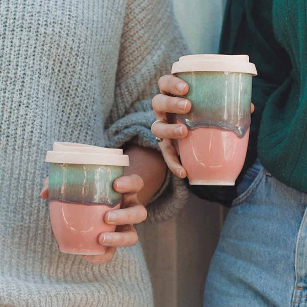 Two people holding pink ceramic travel cup with green and pink gradient glaze and white lids