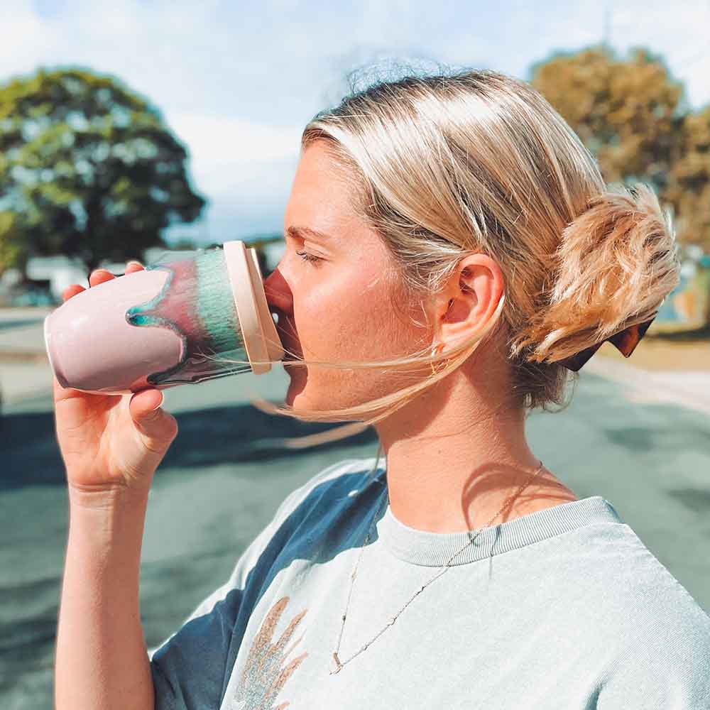 Woman drinking from a pink ceramic travel cup outdoors with trees in the background